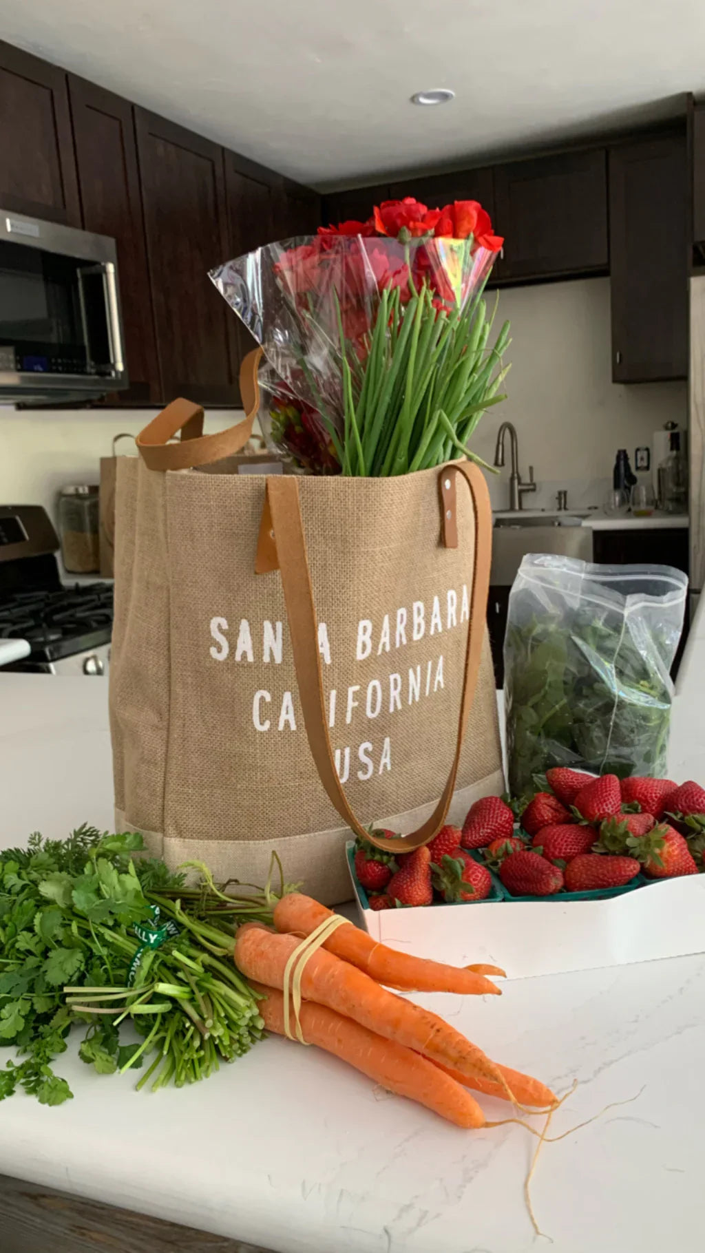 Reusable market tote with fresh carrots, strawberries, greens, and bouquet of red roses on kitchen counter