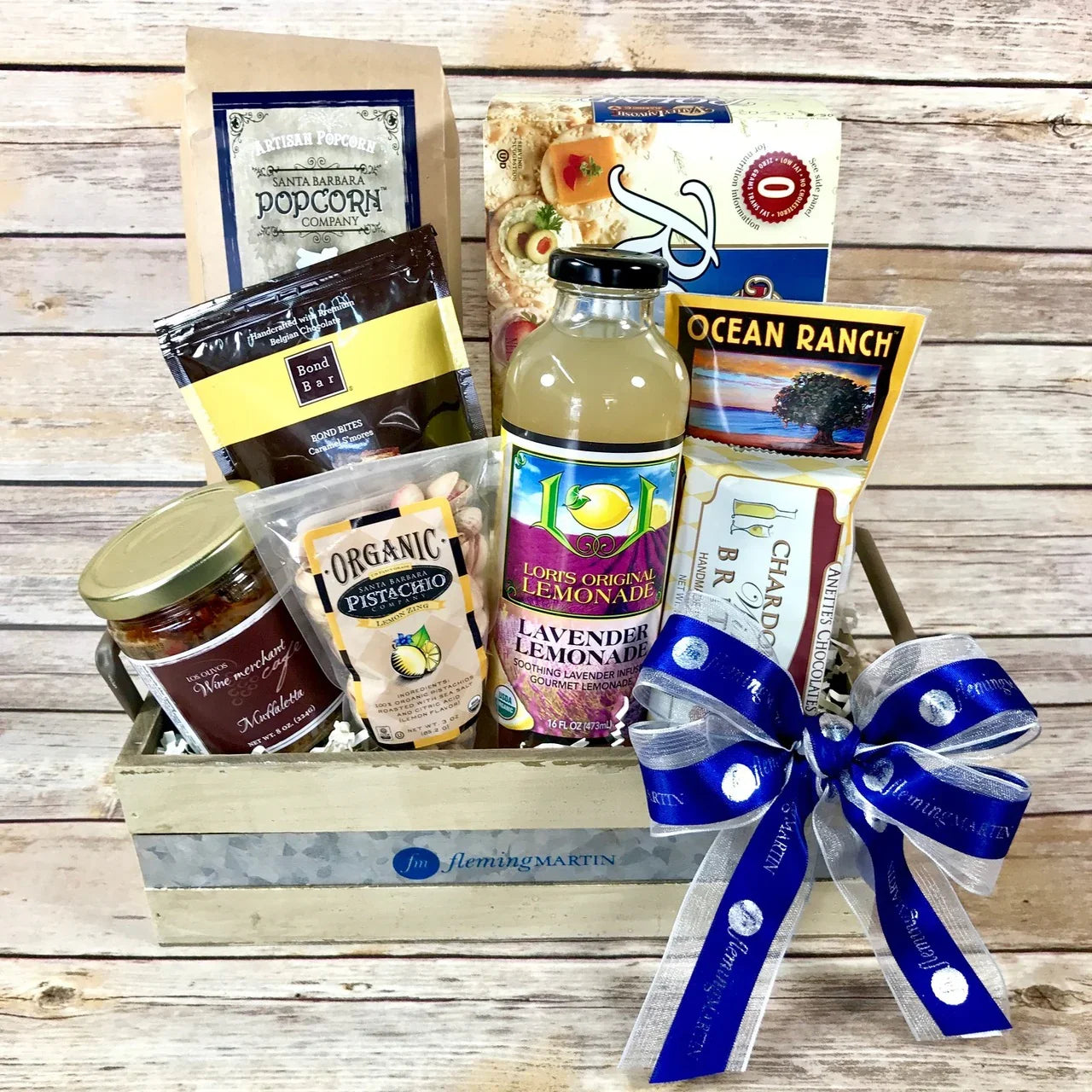 Fleming Martin wooden gift crate with artisanal snacks, lavender lemonade, pistachios, chocolate, and crackers with blue ribbon