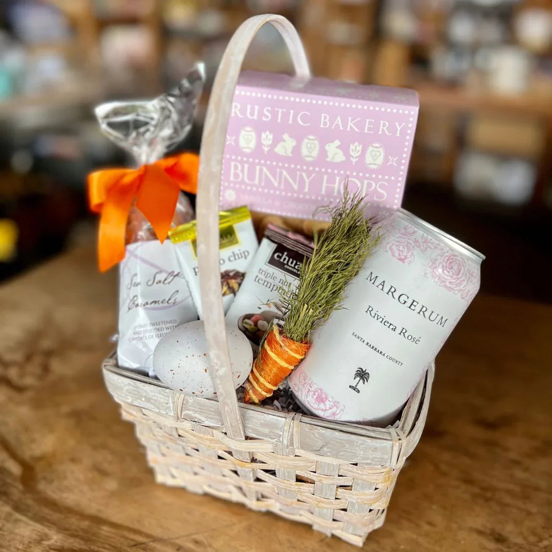 Easter gift basket with Margerum Riviera Rosé can, baked goods, snacks, and decorative carrot on wooden table