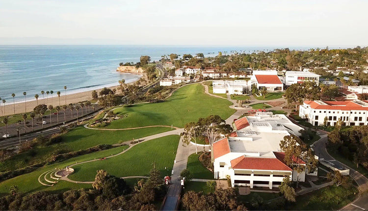 Aerial view of a coastal campus with green lawns, red-roofed buildings, palm trees, and an ocean beach