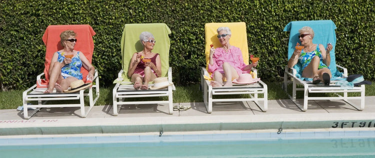 Four elderly women in swimsuits relaxing on colorful poolside loungers with cocktails