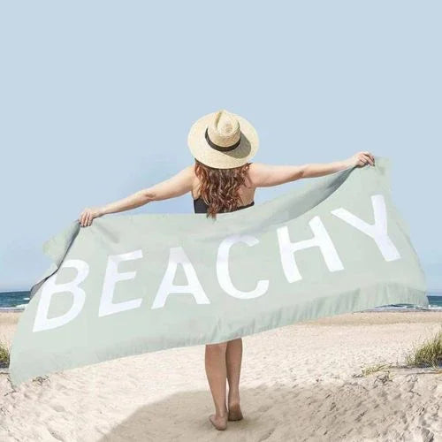 Woman in sun hat holding large green towel with BEACHY text on sandy beach under clear sky