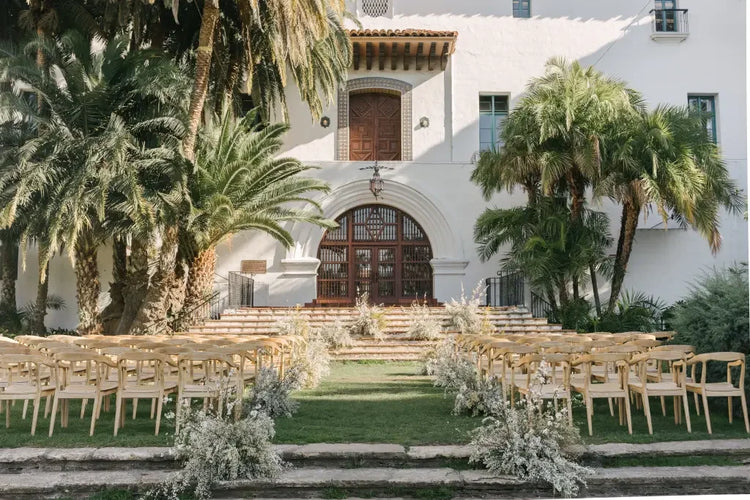 Outdoor wedding setup with wooden chairs and floral decorations in front of a white Spanish-style building with arched wooden doors