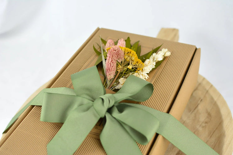 kraft paper gift box with green ribbon and dried pink, yellow, white flowers on wooden surface